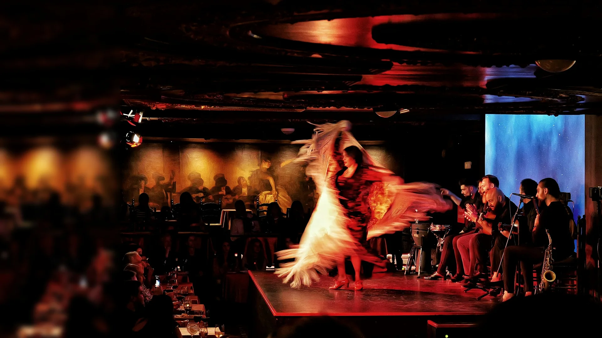 A flamenco dancer in a fringed shawl spins on a raised red stage in a dark Madrid tablao while guitarists and singers sit beside her and diners watch from tables